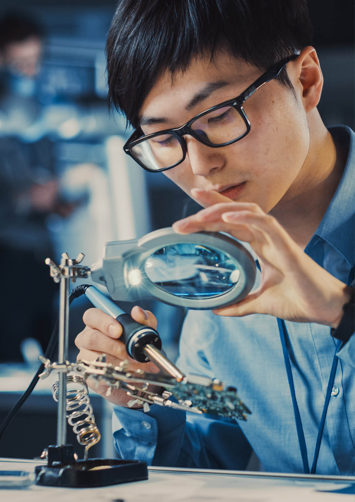 Professional Japanese Electronics Development Engineer in Blue Shirt is Soldering a Circuit Board in a High Tech Research Laboratory with Modern Computer Equipment.