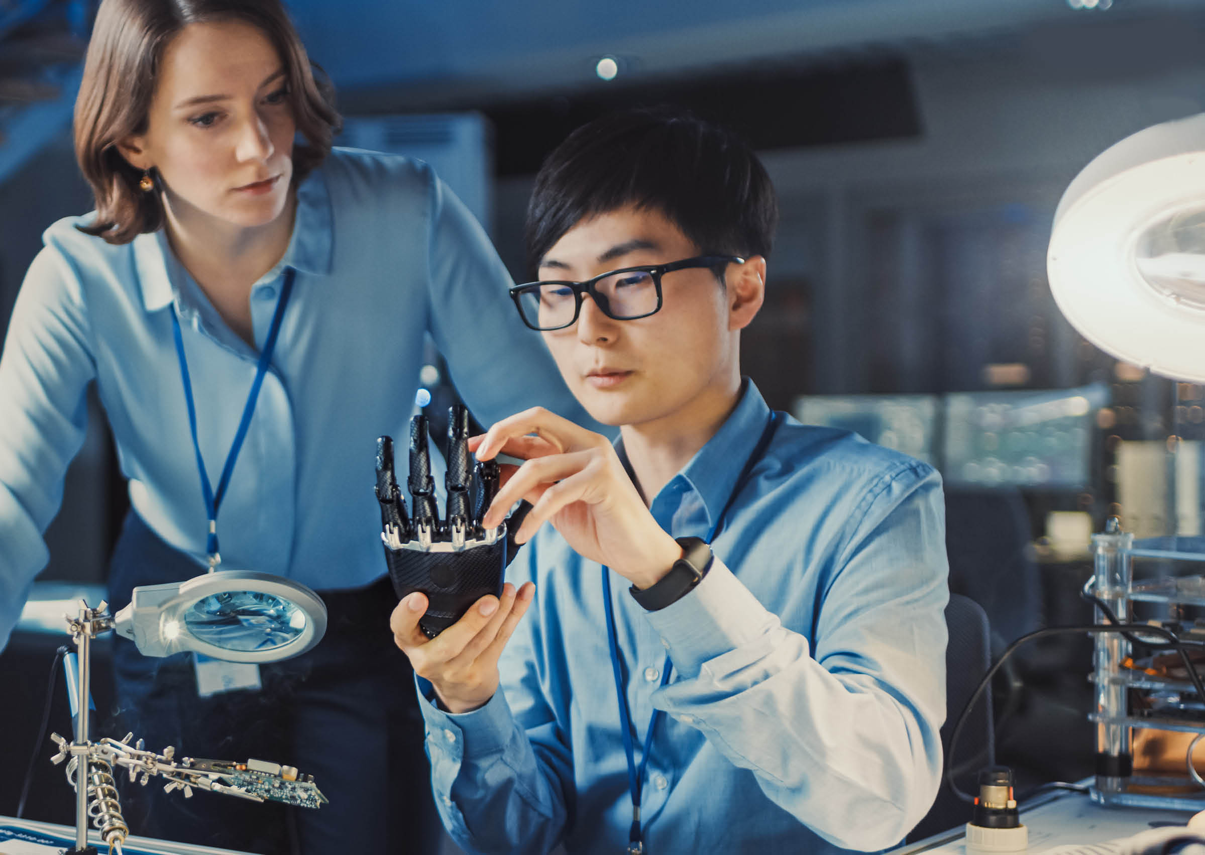 Technological Prosthetic Robot Arm is Tested by Two Professional Development Engineers in High Tech Research Laboratory with Modern Futuristic Equipment. Man and Female Compare Data on a PC