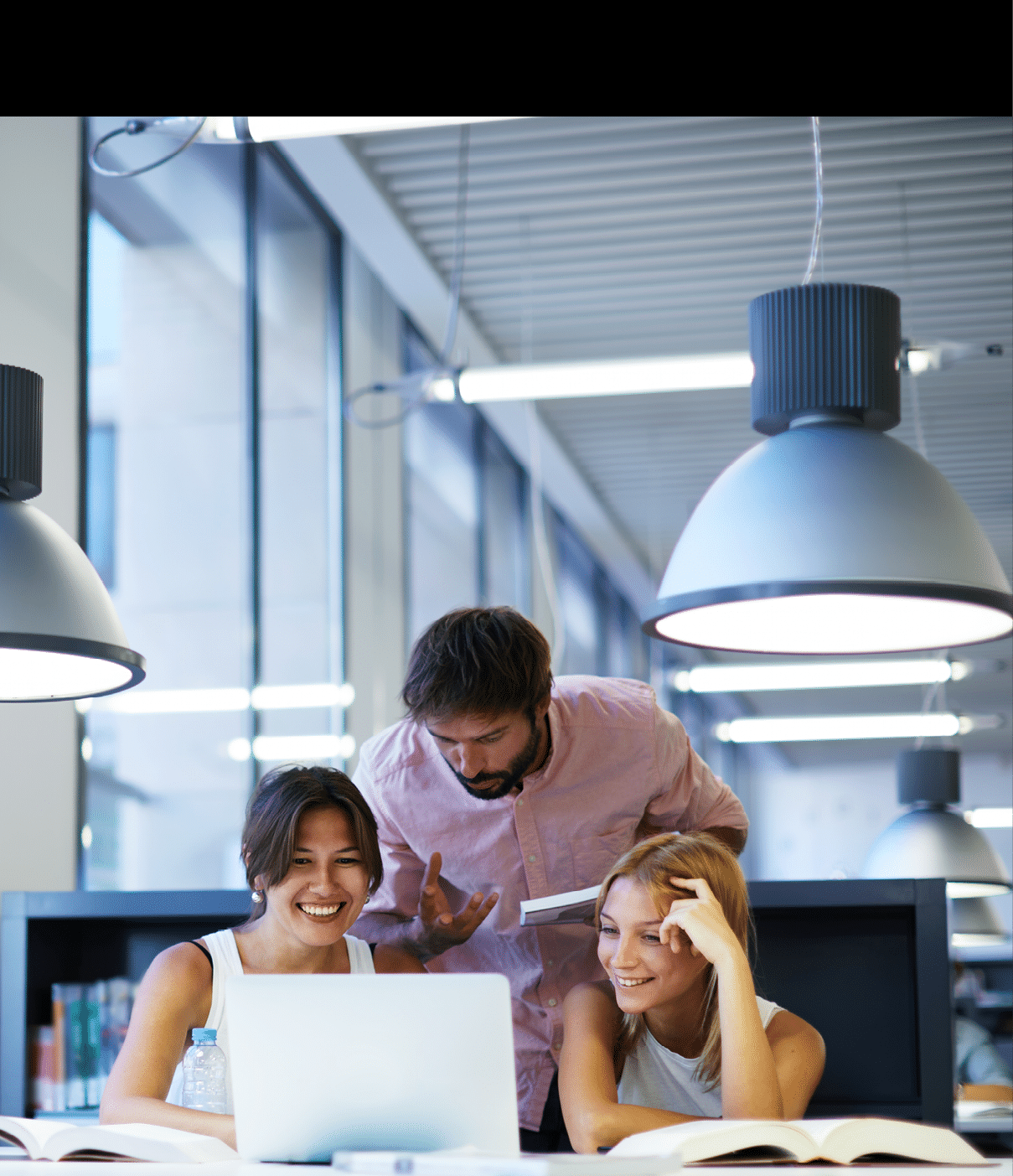Group of international university students having fun studying in library, three colleagues of modern work co-working space talking and smiling while sitting at the desk table with laptop computer