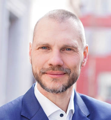 A person wearing a blue suit jacket and white shirt is standing outdoors with a blurred urban background. Description generated by AI