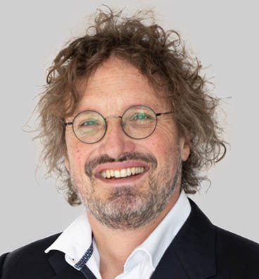 Person with curly hair wearing a white collared shirt and dark blazer against a plain light background. Description generated by AI