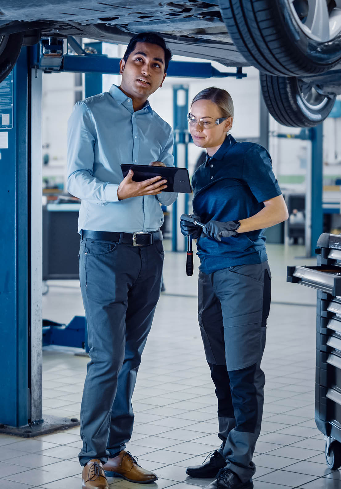 Female Mechanic Talking to a Manager Under a Vehicle in a Car Service. Specialist is Showing Info on a Tablet Computer. Empowering Woman Wearing Gloves and Safety Gloves. Modern Clean Workshop.
