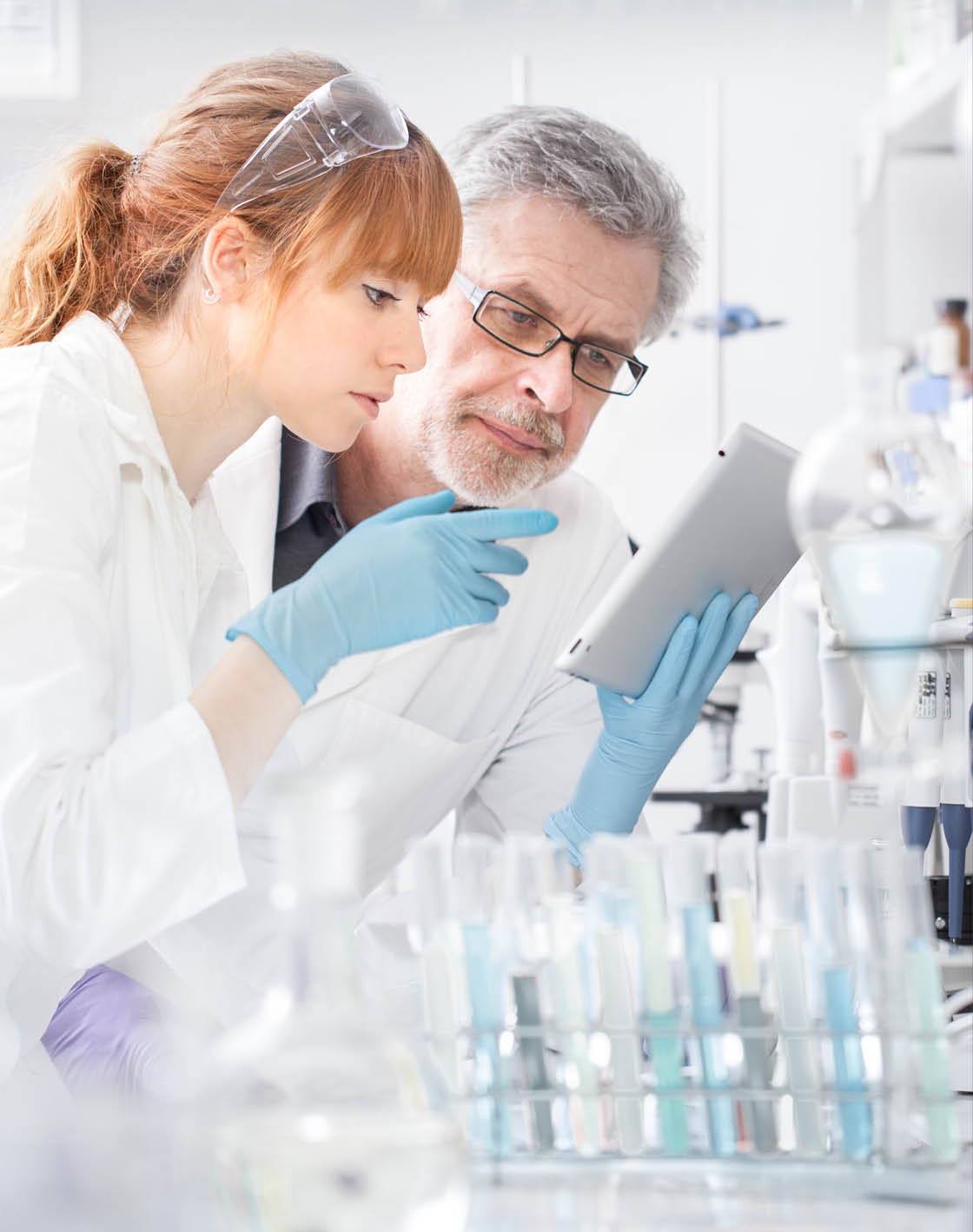 Health care researchers working in life scientific laboratory. Young female research scientist and senior male supervisor looking focused at tablet computer screen evaluating and analyzing study data.