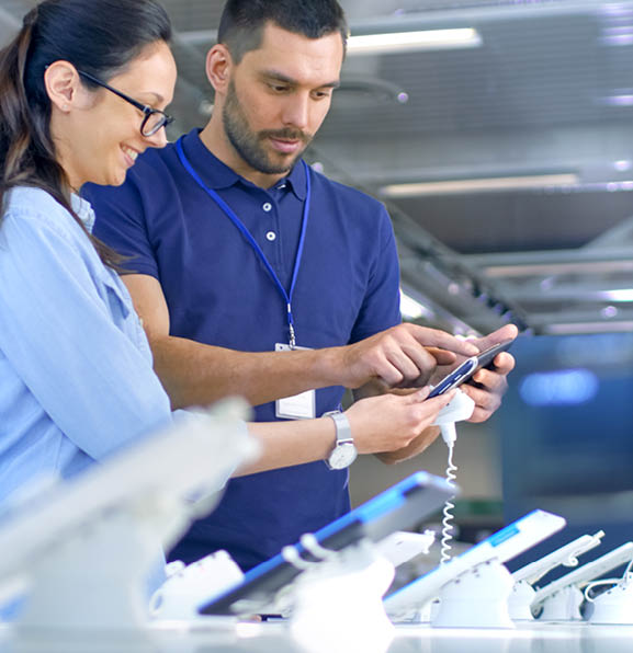 In the Electronics Store Consultant Gives Professional Advice to a Young Woman, She Considers Buying New Tablet Computer and Needs Expert Opinion. Store is Modern, Bright and Has all the New Devices.