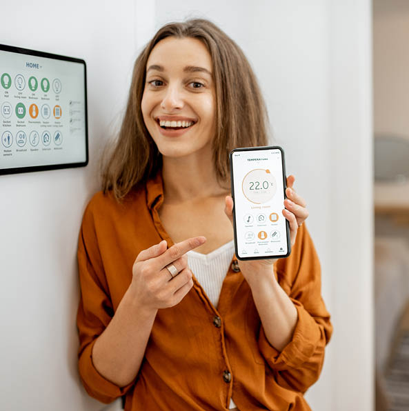 Portrait of a young and happy woman near a touch screen panel for managing smart home. Holding smart phone with a simlpe and convinient mobile application for smart home