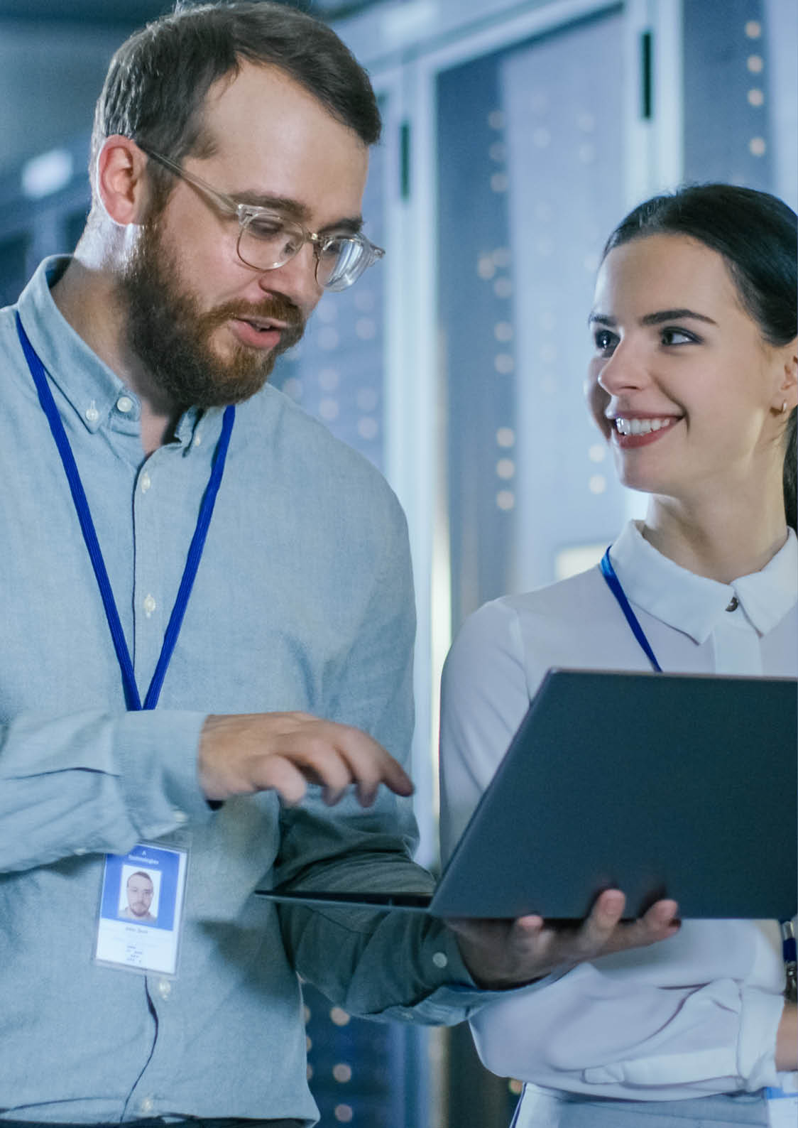 Bearded IT Technician in Glasses and Beautiful Young Engineer Colleague with a Laptop Computer are Talking in Data Center while Working Next to Server Racks. Running Diagnostics or Doing Maintenance 